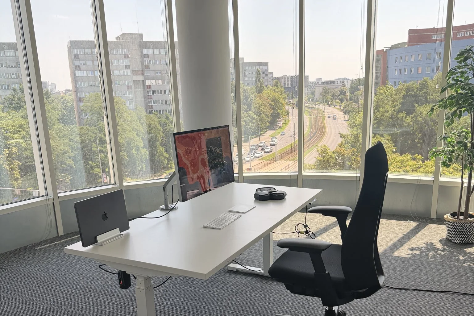 My desk: a single 27-inch monitor, a MacBook in a vertical stand with the lid closed, a keyboard, and a city view through the window.